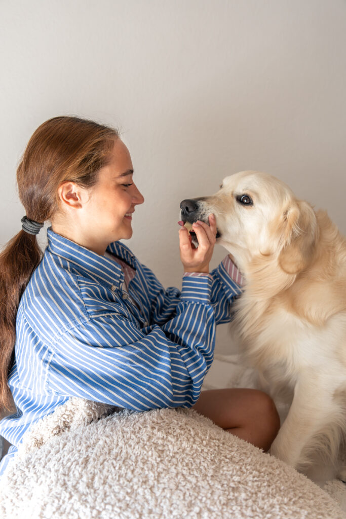 Woman in a striped blue shirt smiles while gently feeding a golden retriever indoors. The scene conveys warmth and companionship.